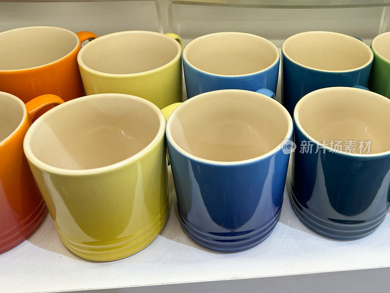 一排彩虹色陶制马克杯的特写图片，展示了红色、橙色、黄色、蓝色和绿色渐变色调的咖啡/茶杯，聚焦于前景，高架视图